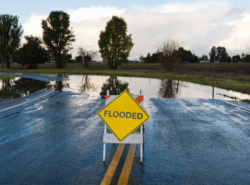 A yellow traffic sign that reads "flooded" is set in front of a road that is completely overrun with water.