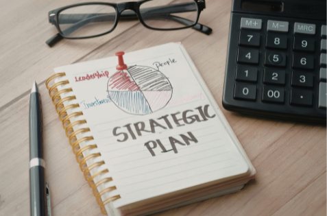 A pen, notebook, calculator, and pair of glasses on a desk. Written in the notebook is "Strategic Plan" with a graph above the words.